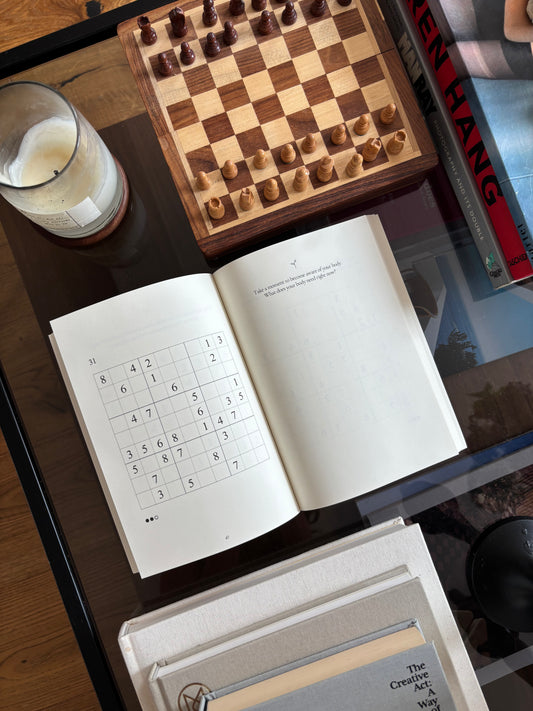Open Mindful Sudoku journal showing a puzzle page, styled on a coffee table beside a chessboard and candle